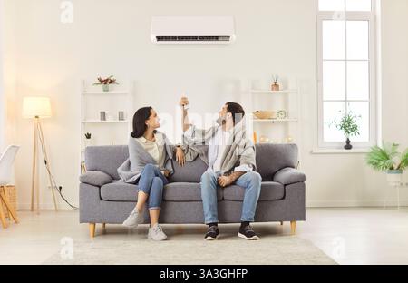 Young couple turning on air conditioner sitting on sofa at home wrapped in a blanket Stock Photo