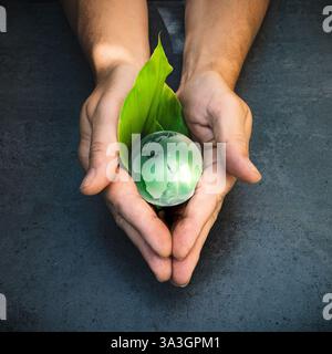 Hands holding a green globe of planet Earth and geeen leaf over a dark grey background Stock Photo