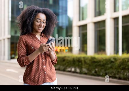 A young woman checks her smartphone in front of an office building, enjoying modern technology and staying connected. Stock Photo