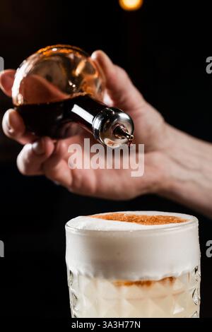 Drop of angostura bitter is falling in Penicillin cocktail. Bartender prepares Penicillin cocktail at the bar. Bartender mixes whiskey, lemon juice, a Stock Photo