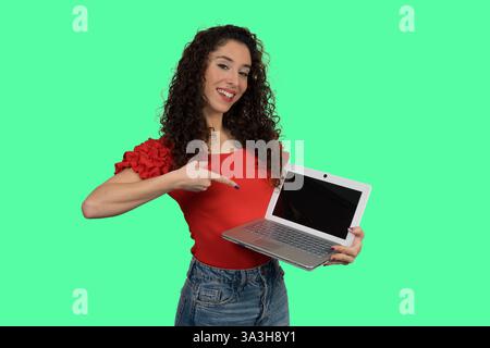 Smiling woman pointing at laptop with blank screen on green background, ideal for mockups and presentations Stock Photo