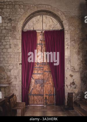 A weathered wooden door with iron details, framed by deep red velvet curtains in an old stone building. The aged walls and warm lighting evoke a sense Stock Photo