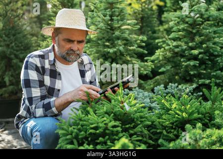 Using digital tablet, sitting. Senior gardener is at work at daytime. Stock Photo