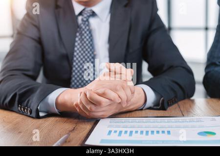 A man in a suit is sitting at a desk with a laptop and a clipboard. He is looking at the clipboard and he is deep in thought Stock Photo