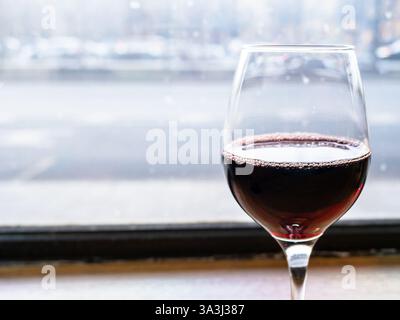 glass of red wine close up on windowsill in bar and view of city street through window in background in winter day Stock Photo