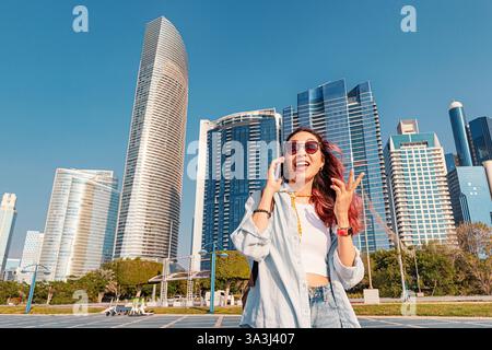 Cheerful young woman with pink hair talking on smartphone in Abu Dhabi, United Arab Emirates, with modern skyscrapers in background Stock Photo