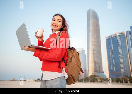 Smiling student with backpack holding laptop and takeaway coffee looking away in Abu Dhabi, United Arab Emirates Stock Photo