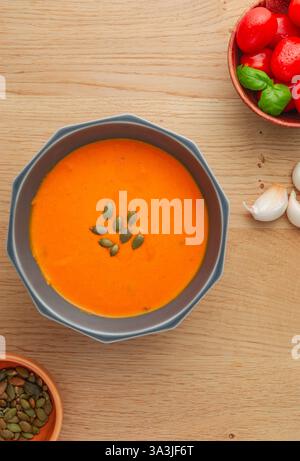 A bowl of soup with a few pumpkin seeds on top sits on a wooden table. There are two bowls of tomatoes and pumpkin seeds on the table as well Stock Photo
