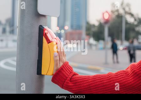 Woman pushing the crosswalk button while waiting to cross the street, observing the red traffic light in a busy urban setting Stock Photo