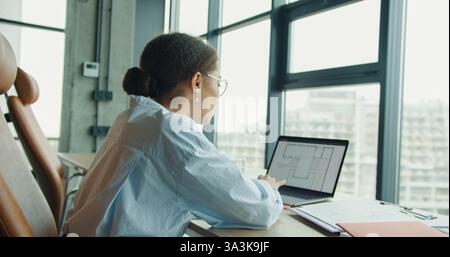 A young professional woman working on a laptop in a bright modern office space, analyzing architectural blueprints on the screen with large windows Stock Photo