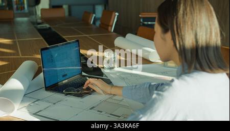 An architect reviewing detailed blueprints on a laptop in a modern conference room, surrounded by rolled plans and sunlight reflecting on a wooden Stock Photo