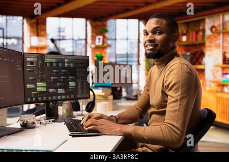 Portrait of smiling IT expert using PC in startup office to design and implementing algorithms enabling machine learning. African american tech venture employee conducts code reviews to fix bugs Stock Photo
