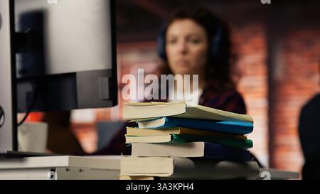 Focus on stack of books on startup office desk with woman in blurred background typing on PC, solving tasks. Pile of books in venture company workspace in front of employee working, camera B closeup Stock Photo