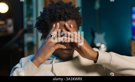 African american man suffering from a migraine and illness, sitting on couch at home and feeling overworked. Concerned young adult struggling with pain, tired and anxious guy. Camera B. Stock Photo