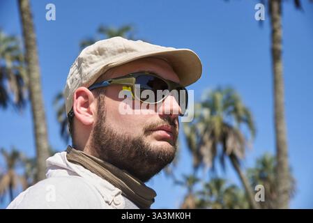 Portrait of a young man with sunglasses and linen beret outdoors on a sunny day, with palm trees in the background. Traveler enjoying nature in El Pal Stock Photo