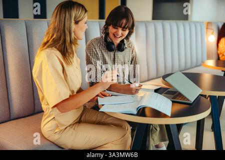 Two joyful college classmates share a moment of laughter while studying together. They are collaborating in a modern lounge, enhancing their learning Stock Photo