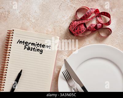 Notepad with intermittent fasting writing, empty plate with cutlery and measuring tape suggesting weight loss through time-restricted eating Stock Photo