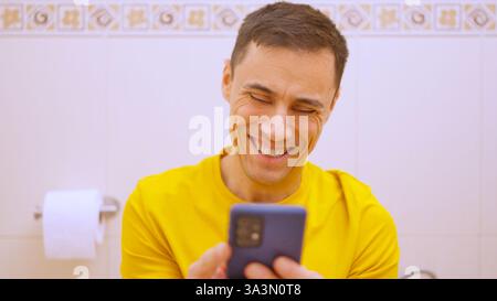 Man laughing while using smartphone on toilet in bathroom Stock Photo