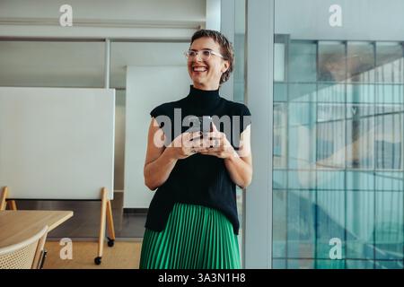 A non-binary entrepreneur smiling confidently while using her smartphone. She stands in a modern office environment, conveying themes of success, inno Stock Photo