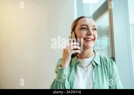 A confident caucasian woman standing by the window, smiling while on a phone call in an office environment. She exudes a sense of professionalism and Stock Photo