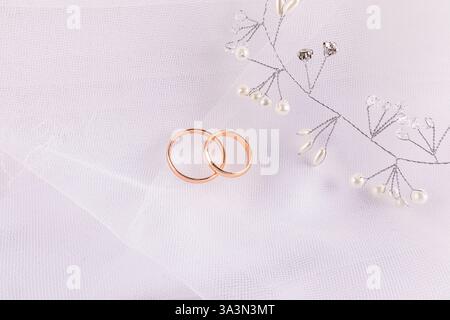 Two classic wedding rings lie on a white chic veil and a satin background. Jewelry made of rhinestones and pearls. The concept of the bride's day Stock Photo