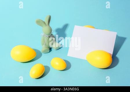 Easter composition with a green velvet bunny, yellow eggs, and a blank white card on a pastel blue background, symbolizing spring celebration and holi Stock Photo