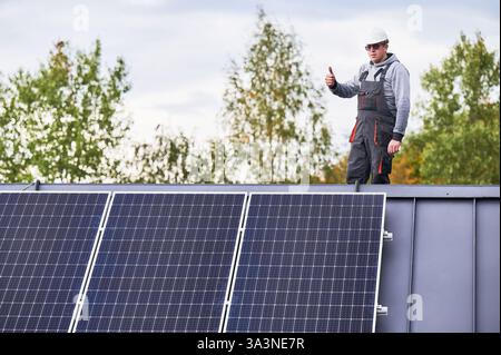 Portrait of man technician installing photovoltaic solar modules on roof of house, showing thumbs up. Electrician building solar panel system outdoors. Concept of alternative and renewable energy. Stock Photo