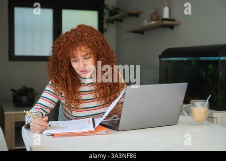 Young woman working from home taking notes and using laptop computer Stock Photo