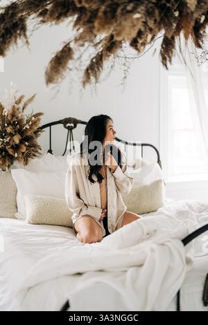 Woman in a satin blouse sitting on a bed in a softly lit bedroom Stock Photo