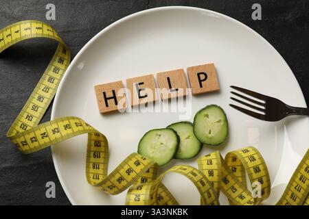 Bulimia. Plate with word Help, pieces of cucumber, measuring tape and fork on dark textured background, top view Stock Photo