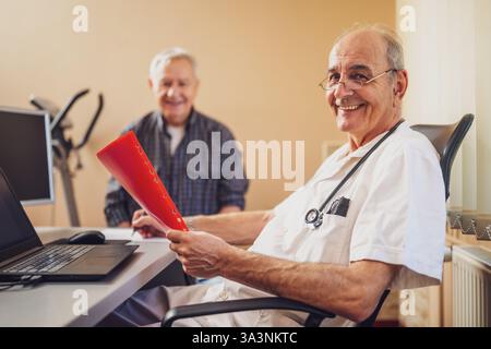 Elderly patient is having exam at cardiologist. Doctor is looking at his medical files. Happy doctor and patient smiling. Stock Photo