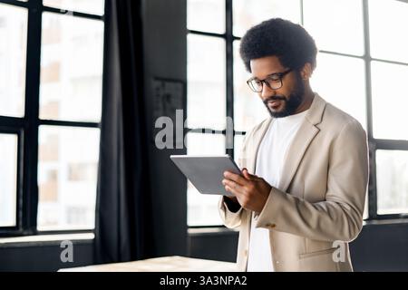 A Brazilian businessman is engrossed in his tablet, standing by a window in a modern loft office, portraying the concept of global connectivity and digital business Stock Photo