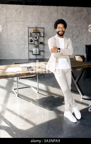 A stylish professional in a beige suit looking at the camera leans casually against an office table, exuding a relaxed yet confident workplace vibe. Full length Stock Photo