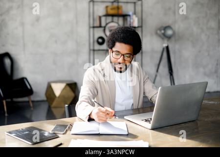 A young African-American businessman in a contemporary office engages with his computer, doing market research and taking notes, busy male employee writing while using laptop Stock Photo