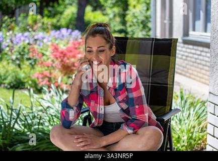 Pretty woman siting comfortably in chair, laughing while talking on phone in colorful garden full of blooming flowers and greenery. Stock Photo