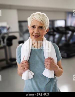 Senior woman with short white hair smiling while wearing blue shirt and towel around her neck, enjoying moment post workout in gym Stock Photo