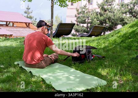 man in red T-shirt sits on green yoga mat in lush grassy park, gently petting black cat inside portable pet carrier. The cat wears red harness, peekin Stock Photo