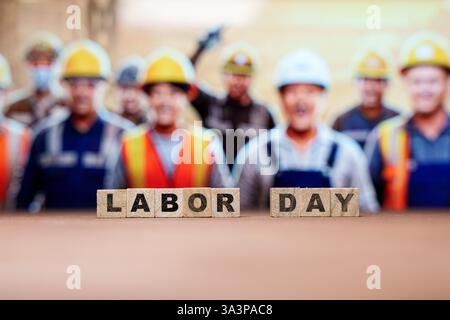 Wooden blocks spelling LABOR DAY placed on a surface, with blurred construction workers wearing safety helmets and vests. Worker appreciation, labor Stock Photo