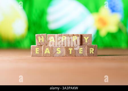 Wooden blocks spelling HAPPY EASTER with colorful Easter eggs and flowers in the blurred background. A festive and cheerful scene symbolizing Easter Stock Photo