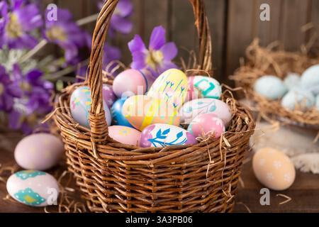 Decorated Easter eggs in a basket with purple daisies on rustic wooden background Stock Photo