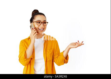 Happy caucasian woman in a yellow shirt and glasses is talking on her smartphone, gesturing with her hand and smiles, standing against an isolated white background. Concept of mobile communication Stock Photo