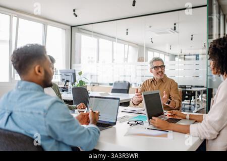 Diverse business team engaged in a brainstorming session in modern office. The senior leader in a brown shirt is passionately explaining his ideas to colleagues who are using laptops and taking notes Stock Photo