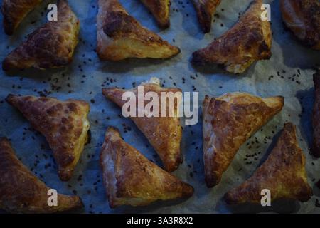 A baking tray filled with freshly baked golden brown triangular cakes. The cakes have a flaky, crunchy texture, are sprinkled with sezam seeds on top. Stock Photo