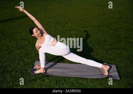 On the yoga mat, exercises. Attractive young fitness woman is on the green grass at daytime. Stock Photo
