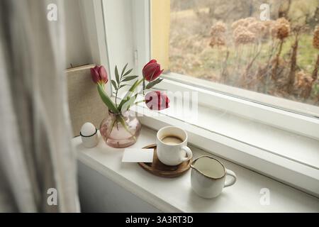 Easter spring still life. Cup of coffee, floral bouquet on window sill. Red tulips flowers, olive branches in vase. Milk pitcher, egg. Business card Stock Photo