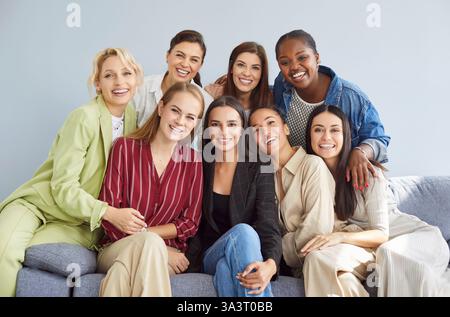 Portrait of happy young diverse women group looking at camera sitting on sofa on gray background. Stock Photo