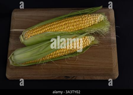 Fresh organic corn on the cob with green husks partially peeled, placed on a rustic wooden cutting board against a dark background Stock Photo