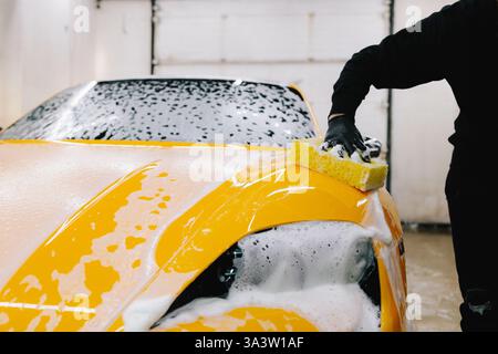 Man washes the sports car using a soapy solution and sponge before detailing procedure. Meticulous car detailing concept at the car service Stock Photo