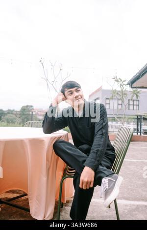 A young man in casual clothing sits at an outdoor table, lost in thought. He rests his head on his hand, looking away with a thoughtful expression. Th Stock Photo