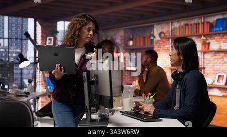 Startup intern holding laptop asks questions to senior technician coding advanced AI algorithm. Employee requests help from CTO manager in Silicon Valley workplace, camera B Stock Photo